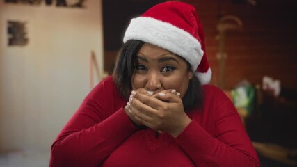 Woman covering mouth in surprise wearing santa hat on construction site with blurred indoor background. - Powered by Adobe