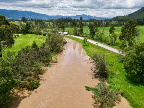 Aerial drone view directly above the swollen, muddy Río Bogotá as it flows through flat green fields and rural landscape near Zipaquirá, Cundinamarca, Colombia.