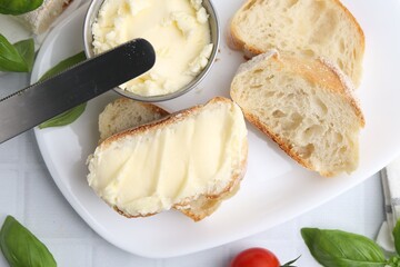 Slices of baguette with butter, basil and knife on white tiled table, flat lay