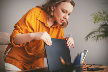 Blonde business woman in bright orange shirt working diligently at home office on laptop and tablet during remote freelance project