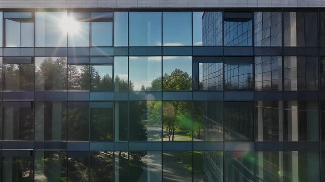 Bright sun flare reflecting on the geometric glass facade of a modern corporate building with park reflections