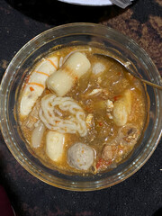 A close-up overhead view of a spicy Asian soup in a clear glass bowl, featuring noodles, meatballs, and fish cakes.