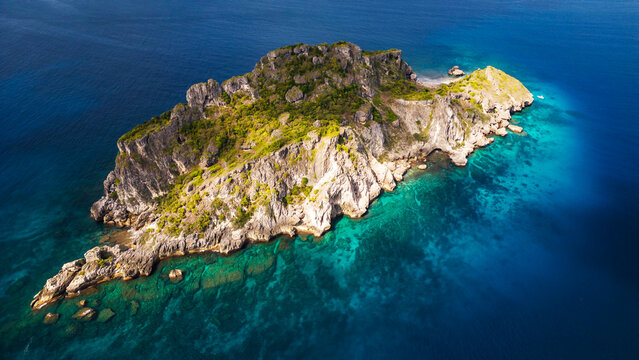 A dramatic, high-angle aerial view of a small, steep, rocky tropical island covered with green vegetation, surrounded by deep blue and clear turquoise water. - Powered by Adobe