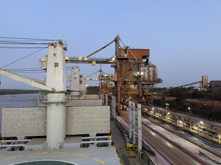 Massive port crane unloads petroleum coke with grab bucket onto conveyor system at modern aluminum production terminal. 443