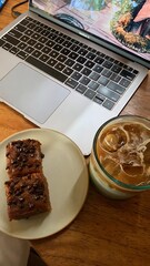 A cozy workspace setup with a laptop, iced coffee, and chocolate chip cake slices on a wooden table — perfect for a relaxing work or study session.