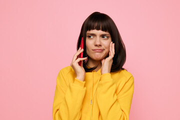 A young woman in a bright yellow hoodie stands against a pink backdrop, talking on a phone with a puzzled expression, conveying stress, doubt, or a thoughtful conversation.