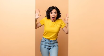 Shocked woman with hands up in surprise wearing a yellow shirt against a peach colored studio backdrop
