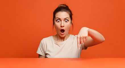 Shocked woman pointing down with an open mouth against a vibrant orange background in studio shot