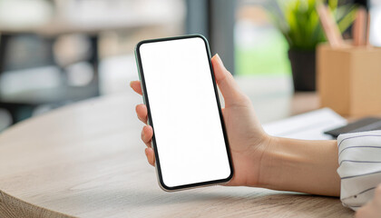 Close-up of a woman's hand holding a vertical smartphone with a blank white screen over a light wooden table in an office/cafe.
