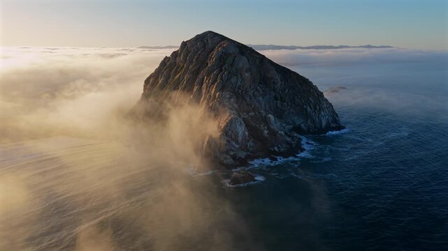 Breathtaking and captivating aerial view showcases Morro Rock island prominently rising from mysterious foggy waters, beautifully bathed in the warm glow of a stunning sunrise. Morro Bay, California
