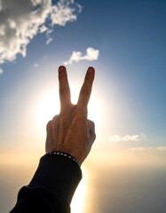 A hand makes the peace sign against a sky with sun rays and soft clouds