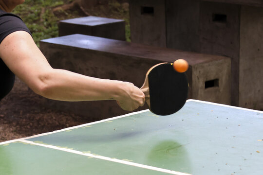 Focused hand holding black paddle, playing game of table tennis outdoors. An orange ball hitting racket during fun sport activity on green table in park
