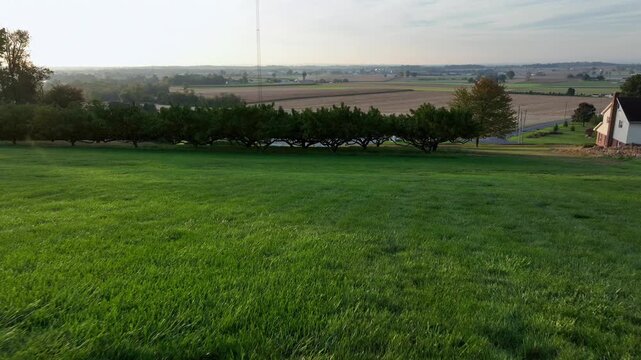 Hills grass in garden of American farmstead house in USA. Sunset time in fall season. Farmland fields with cultivation in the valley of town. Wide shot. Drone forward shot.