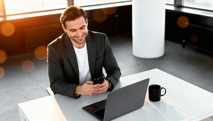 Man in suit jacket sitting at desk using smartphone and laptop.