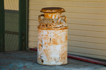 A rusty vintage milk can sits on a wooden porch against a beige wall the metal container shows signs of age and weathering evoking a sense of nostalgia and rural history