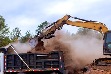 An excavator is loading dirt into truck while dust debris fill air at busy construction location.