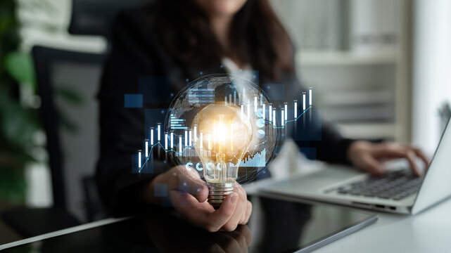 A business professional showcases a light bulb symbolizing innovation while surrounded by digital graphs and charts in a modern office setting. Scalp - Powered by Adobe