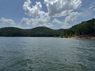 Sunny lake view with forested hills and rippling water under scattered clouds
