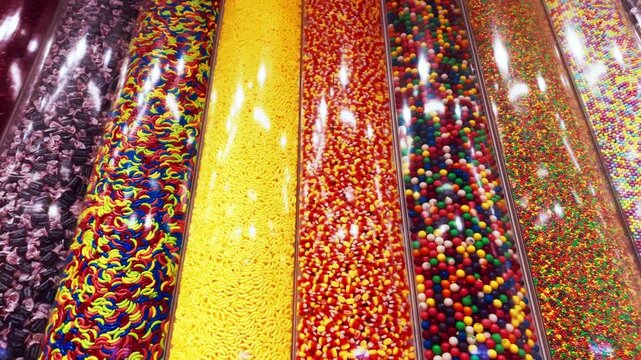 Close-up POV of colorful candy dispensers filled with assorted sweets gummy worms, sprinkles, small chocolate pieces, and rainbow candy beads.