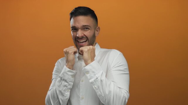 Young hispanic man in a white shirt displays fear against an orange background, hands clenched near his mouth, embodying intense emotion and anxiety.