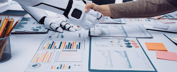 A human and a robot shaking hands in a modern office setting, symbolizing collaboration and innovation, with data charts and tools laid out on the table.Noogenesis