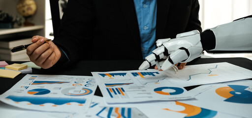 A businessman collaborates with a robotic arm to analyze financial reports, demonstrating the fusion of technology and human expertise in modern business environments.Noogenesis