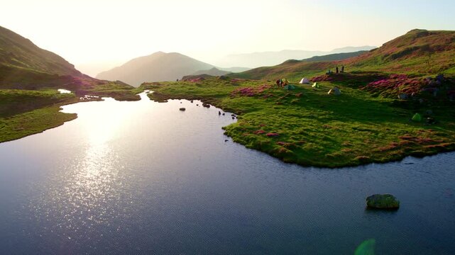 Aerial shot gliding over, still alpine lake reflecting a bright sun flare. Colorful tents are pitched on the green, flower-dotted shore, surrounded by mountain peaks in the hazy, early morning light.
