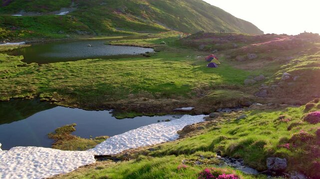Aerial photo showing a colorful campsite next to a small alpine lake in Rodnei mountains. Green grass and pink flowers, patches of white snow, illuminated by a ray of sunlight