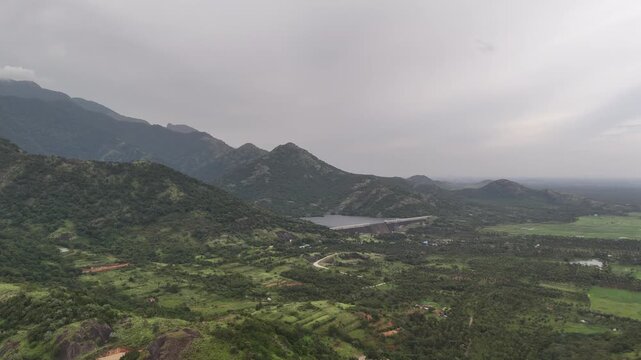 A wide aerial video of a large concrete dam holding back a reservoir, nestled at the base of a majestic, green mountain range. The surrounding landscape includes lush valleys, forests, and small rural