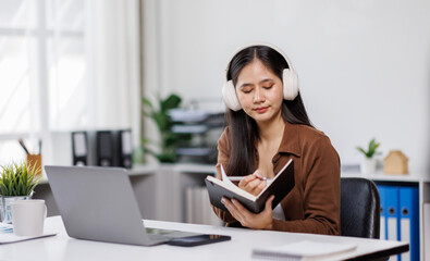 Smiling woman takes notes at her office, wearing headphones listening music at work using laptop remote work. Online education concept

