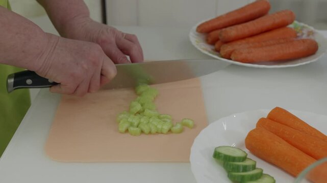 Professional chef chopping fresh celery with precision on wooden cutting board, surrounded by raw carrots and cucumber slices in kitchen setting