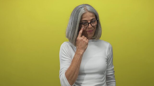 Woman with grey hair points index finger to eyeglasses in studio with yellow backdrop; playful curiosity.