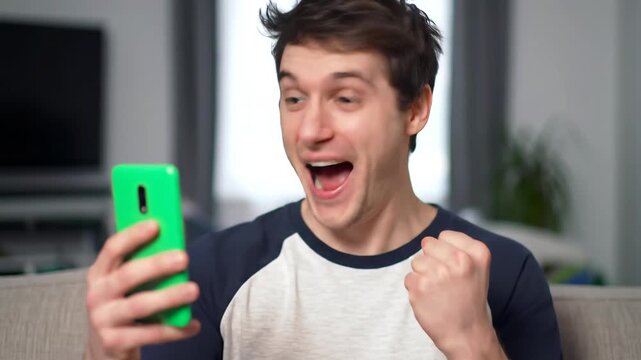 Excited young man celebrates a victory or good news while looking at his smartphone expressing joy and success with a fist pump gesture in a home setting.