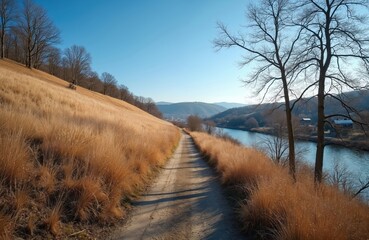Dirt path leads beside dry golden grass and blue river through valley. Bare trees line hill under clear sky. Rural landscape with distant town.