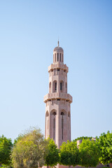 Fototapeta premium A tall Islamic minaret tower rising above lush green trees under a bright, cloudless blue sky in Oman. The tower features elegant arches, beige stonework, and traditional Middle Eastern architecture