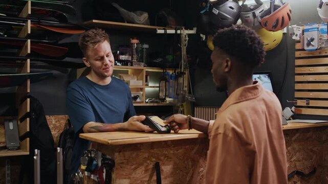Young Black man paying for service in modern repair shop with credit card, young Caucasian worker holding pos terminal