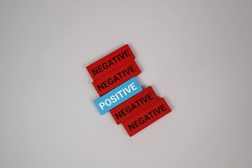A diagonal stack of colored blocks shows a single blue 'POSITIVE' tile among red 'NEGATIVE' tiles against a vivid white backdrop, illustrating the contrast between optimism and pessimism.