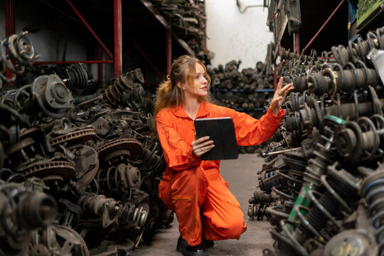 Female technician worker checking inventory with tablet device in garage scrap yard old car part workshop warehouse.