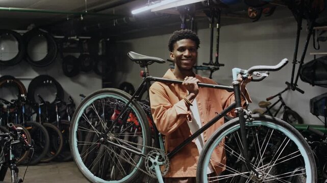 Young African American man holding bicycle standing in bike repair shop, looking at camera, medium portrait