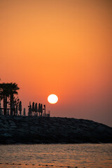 Fototapeta premium A dramatic, intense orange and red sunset or sunrise over the sea, silhouetting beach umbrellas and palm trees on a rocky shoreline