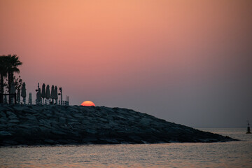 The sun setting directly behind a dark, rocky breakwater, silhouetting palm trees and folded beach umbrellas against a deep orange, hazy sky over the Arabian Gulf