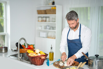Cook chops vegetables in kitchen. Chef in apron prepares healthy meal in modern kitchen. Cook with vegetables in white kitchen. Cook prepares breakfast with veggies at home kitchen.