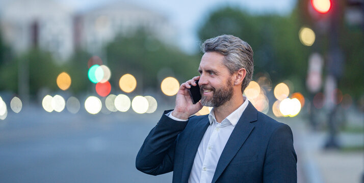 Middle aged business man talking on a smartphone while walking in the city at night. Business late call. Business conversation overtime work. Business man man in a suit speaking on the phone outdoor.