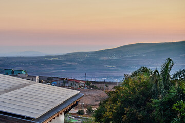 Mountain valley landscape during dusk with red roof houses, fields and layered hills stretching into the misty distance.