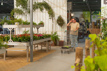 Man shopping for plants and flowers inside a garden center, pushing a trolley with potting soil bag