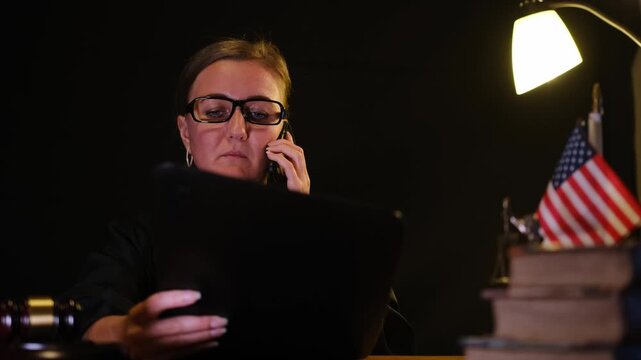 Female judge in black robe speaking on the phone and checking a document at her desk with books