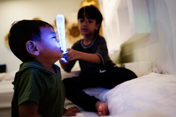 A young boy stands on the bed holding two glowing toys, enjoying imaginative play in a dim home...