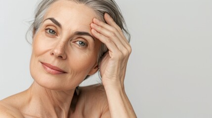 Obraz premium A senior Caucasian woman with gray hair gently touches her forehead. She has a calm expression and smooth skin, set against a neutral background.