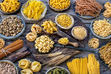 Different types of uncooked pasta on rustic wooden table, in the spoons, cutting board and bowls 