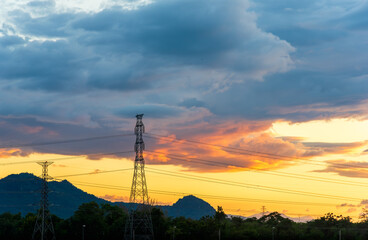 Obraz premium Silhouette of high-voltage electricity pylons transmission towers against a dramatic, cloudy sunset sky with an orange glow over a mountain landscape.
